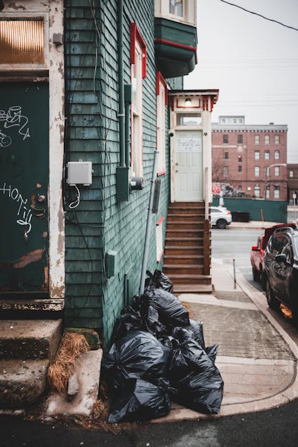 A row of black garbage bags filled with waste are loosely stacked at the base of a green wooden building, positioned on a concrete sidewalk next to a small set of wooden stairs leading to a white door with red trim. The building’s exterior features green shingles with a textured surface, and adjacent to the stacked rubbish, there is a weathered metal utility box mounted on the wall, along with exposed electrical wires. To the left, an entrance door with graffiti tags and a weathered surface is visible, with concrete steps leading into the building. In the background, a parking area with cars and a large multi-story brick building can be seen under a cloudy sky. The scene appears to be in an urban environment, where independent waste disposal methods might be utilized for rubbish removal services provided by Rubbish Removal Knightsbridge, reflecting typical private waste handling practices in this area.
