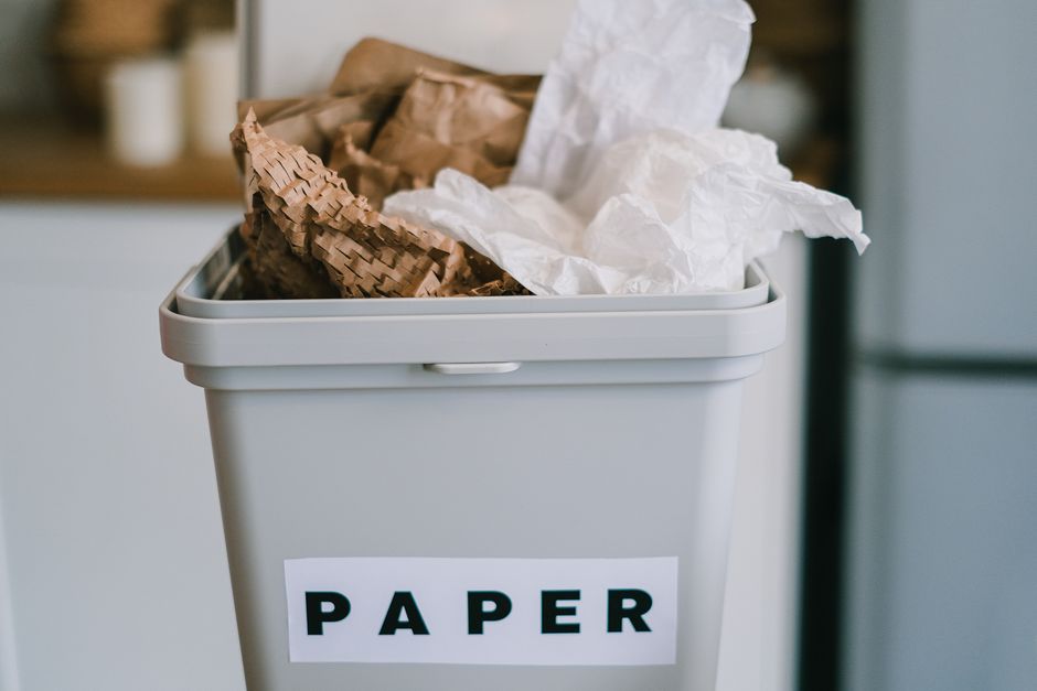 A white rectangular bin with a slightly rounded top edge and a smooth, matte finish is positioned in an indoor environment, likely a kitchen or office space. The bin is partially filled with crumpled paper waste, including both white and brown paper, with some pieces spilling over the rim. The brown paper appears to be packaging material with a textured, slightly rippled surface, while the white paper is crumpled and irregular. A label with the word 'PAPER' in bold black letters is affixed to the front of the bin, indicating its designated use for paper waste. The background is softly blurred, but a dark-colored appliance or piece of furniture is faintly visible, contributing to a domestic or commercial indoor setting. Lighting in the scene is natural and diffuse, creating gentle shadows and highlights that emphasize the textures of the paper and the smooth surface of the bin. This image aligns with waste management practices involving the separation and collection of recyclable paper, natural to services provided by companies like Rubbish Removal Knightsbridge in managing non-technical, on-site waste disposal tasks.