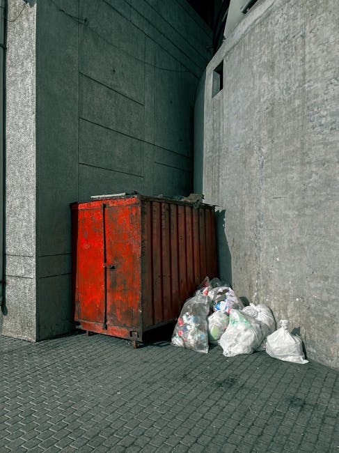 A red metal waste skip with visible rust and chipped paint is positioned against a dark green wall and a light grey concrete wall in an urban alleyway. The skip's surface appears weathered with signs of corrosion and peeling paint. Adjacent to the skip, on the paved ground, are several clear plastic bags filled with mixed rubbish, including paper, plastic bottles, and packaging materials. The ground is covered with dark, textured paving stones, forming a flat surface. The alleyway is enclosed by the building walls, with a small, dark opening or window visible high on the light grey wall on the right side. The lighting is natural, creating soft shadows on the surface of the skip and the pavement, emphasizing the textures of the materials. This scene illustrates a typical private waste collection area, often managed by services such as Rubbish Removal Knightsbridge, offering an alternative to council rubbish disposal methods for immediate and discreet waste removal near commercial or residential premises.