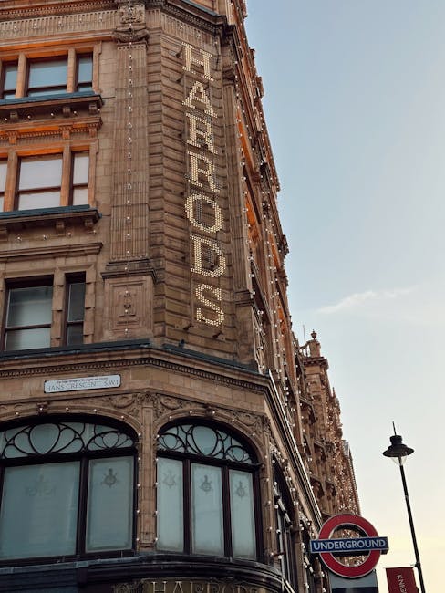 A wide street scene in an urban area with historical and modern architecture. On the left, there are tall, ornate brick buildings with bay windows, decorative facades, and turrets, typical of late Victorian or Edwardian style. The ground level features retail storefronts with large glass windows, one of which displays a sign for a jewelry store. In the center, a prominent historic building with intricate stonework, arched windows, and a domed roof with decorative elements is visible, with a large billboard partially obscuring part of its facade. To the right of this building, a curved glass awning extends from a modern shopfront, with pedestrians walking along the sidewalk beneath it. The street is lined with multiple parked cars and some moving vehicles, including a distinctive red double-decker bus that is characteristic of London. People, some dressed in business attire and others in casual clothing, are walking or standing, and the sky above is a gradient of pale blue with a few scattered clouds, suggesting early evening or late afternoon. This scene, part of the area around Harrods, reflects city street life with an emphasis on private waste collection and rubbish removal services in a busy commercial district.