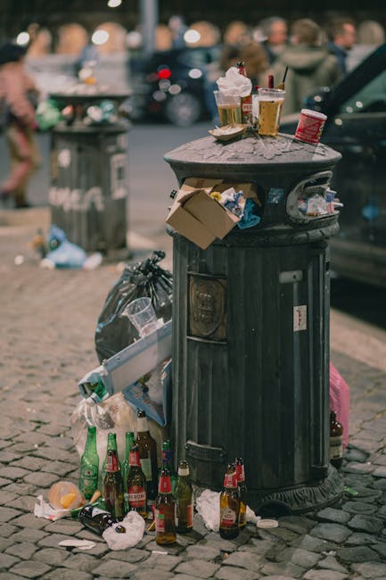 A black, cylindrical public rubbish bin with a textured surface and a rounded, flat top located on a cobblestone pavement in an urban setting. The top of the bin is cluttered with used paper cups, a glass of beer with a frothy head, and a small red food container. Several discarded bottles, mostly glass, including green and brown beer bottles, are scattered on the ground around the base of the bin. On the side of the bin, there is an open slot with a partially filled plastic trash bag hanging out, along with a cardboard box and additional waste materials. In the background, there are blurred figures of pedestrians, parked cars, and another grey waste container. The overall scene depicts a littered street environment, with visible signs of improper waste disposal that could relate to the importance of responsible rubbish removal and alternative waste handling methods, as promoted by local services like Rubbish Removal Knightsbridge.
