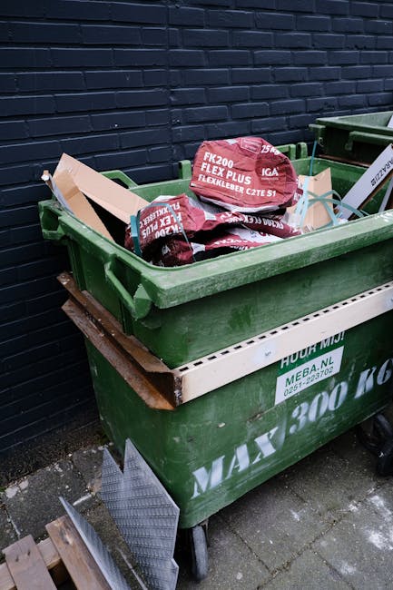 A large green plastic rubbish dumpster filled with torn red and black bags of garden or lawn waste, situated outdoors against a dark brick wall. The bags, branded with text, appear to contain organic material, and the dumpster is mounted on four wheels for mobility. Below the green container, there is a white horizontal bar with additional text, and part of a wooden platform or pallet can be seen supporting the bin. The surrounding ground is paved, with some dirt or debris nearby. This scene reflects a typical outdoor waste collection setup where private rubbish removal services, such as those by Rubbish Removal Knightsbridge, might handle garden or yard waste outside a residential or commercial property, providing an alternative to council collection or on-site clearance.