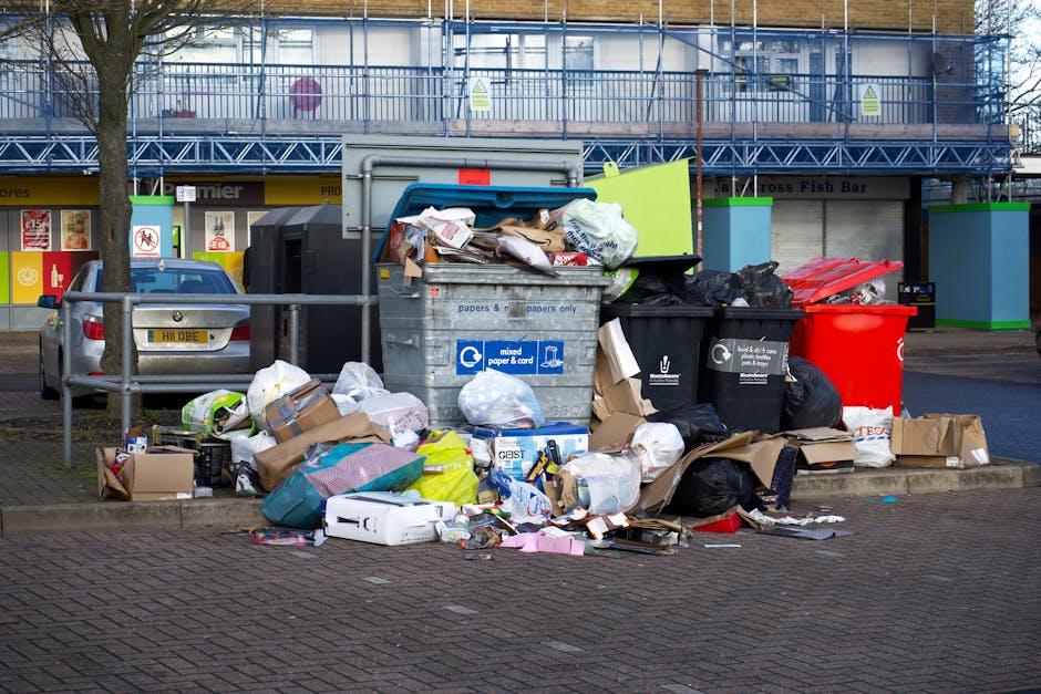 A black, cylindrical public rubbish bin with a textured surface and a rounded, flat top located on a cobblestone pavement in an urban setting. The top of the bin is cluttered with used paper cups, a glass of beer with a frothy head, and a small red food container. Several discarded bottles, mostly glass, including green and brown beer bottles, are scattered on the ground around the base of the bin. On the side of the bin, there is an open slot with a partially filled plastic trash bag hanging out, along with a cardboard box and additional waste materials. In the background, there are blurred figures of pedestrians, parked cars, and another grey waste container. The overall scene depicts a littered street environment, with visible signs of improper waste disposal that could relate to the importance of responsible rubbish removal and alternative waste handling methods, as promoted by local services like Rubbish Removal Knightsbridge.