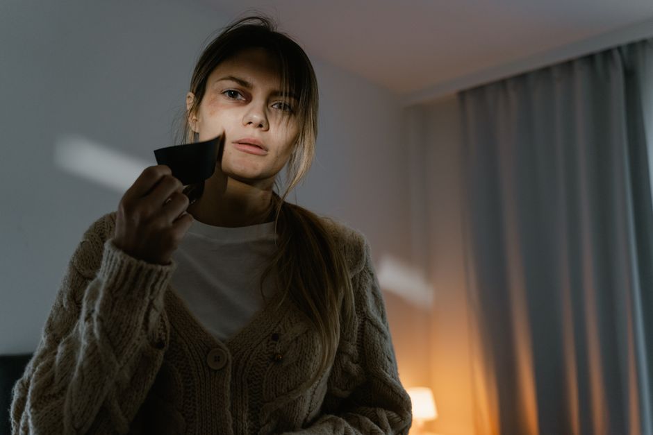A young woman with light skin and long brown hair tied back, wearing a beige knitted cardigan over a white top, holds a black telephone receiver close to her face while looking directly at the camera with a serious expression. She is situated in a modern interior space with soft, ambient lighting, a blue curtain hanging on the right side, and a warm light source illuminating the background. The setting appears to be a living room or bedroom. This scene, while primarily personal, subtly reflects the context of alternative or private communications, which can be related to independent rubbish management services, such as those provided by Rubbish Removal Knightsbridge, working outside standard municipal disposal channels.