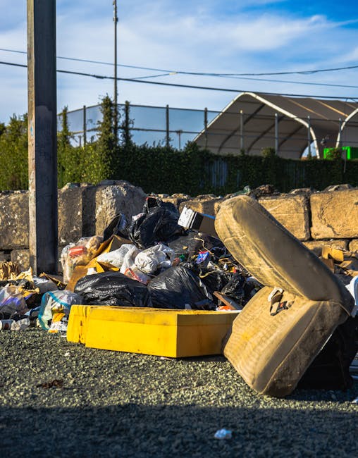 A pile of mixed household rubbish and waste materials is spilled across a rough asphalt surface in an outdoor setting. The debris includes black plastic garbage bags, discarded packaging, and various small waste items, with some of the rubbish spilling out of a yellow plastic container that lies on its side. Nearby, an old, dirty, beige upholstered armchair with visible fabric wear and stains is partially stacked among the rubbish, leaning against a low stone wall constructed from large, uneven grey stones. In the background, there is a tall metal pole with some electrical wires stretching across the scene, along with a fence and green shrubbery. Behind the fence, a large, curved, semi-transparent industrial canopy structure is visible, set against a bright blue sky with white clouds. The lighting indicates daytime with natural sunlight illuminating the scene, emphasizing the textures of the trash, chair, and surrounding environment. Although the scene focuses on waste, it subtly illustrates the need for proper rubbish collection and disposal, aligning with services like those offered by Rubbish Removal Knightsbridge for alternative waste handling solutions.