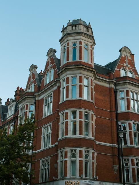 A historic multi-story red brick building with intricate white window frames and decorative architectural details, situated at the corner of a street. The structure features a prominent cylindrical turret topped with a small domed roof, and multiple gabled sections with ornate cornices. The building's facade includes large, vertical sash windows, some with rounded tops, and is set against a clear, pale blue sky. In the foreground, a tall black street lamp is visible, and part of a green leafy tree extends into the scene's lower left corner. The setting suggests an urban environment with careful preservation of traditional architecture, which may relate to private property or buildings associated with local council regulations concerning waste management, as seen through the context of rubbish removal services provided by Rubbish Removal Knightsbridge.