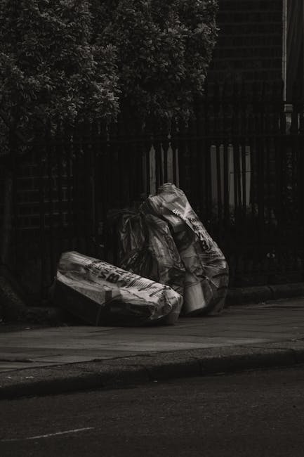 Two large black garbage bags filled with waste material are positioned on the pavement near a black metal fence with vertical bars. The bags appear to contain bulky refuse, possibly cardboard or plastic items, and are crumpled and tied at the top. The surrounding area includes a patch of greenery with dense foliage visible behind the fence, and a section of a building or structure partially seen to the right. The scene is set outdoors in natural light, with the bags placed on a street or roadway, indicating a collection point for rubbish removal. The overall scene suggests an informal waste disposal situation, typical of private rubbish collection services like those provided by Rubbish Removal Knightsbridge, aligning with the context of alternative waste handling and on-site clearance in urban settings.