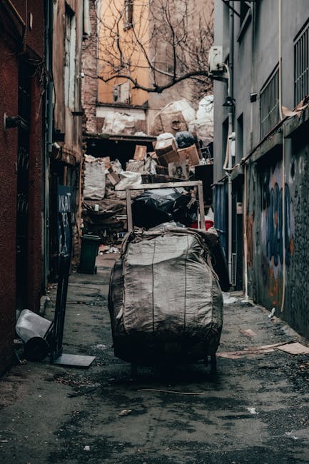 A narrow urban alleyway with rough, uneven pavement and darkened patches, cluttered with various discarded items. In the foreground, a large, grey, weathered sack or bag is positioned centrally, filled with unidentified waste materials. Behind it, there is an accumulation of cardboard boxes, black plastic bin liners, and miscellaneous rubbish piled haphazardly against a chain-link fence and a brick wall. The surrounding structures consist of aged brick and weathered siding, with graffiti visible on some surfaces, indicating a neglected or industrial area. Overhead, bare tree branches extend into the scene, suggesting a cold or dormant season. The alley is dimly lit, with soft, natural light diffusing through the narrow space, highlighting the textures of the debris and urban environment. The scene appears to involve an independent rubbish collection in an alleyway, consistent with the services of Rubbish Removal Knightsbridge or similar waste management providers, emphasizing on-site clearance or alternative waste handling methods.