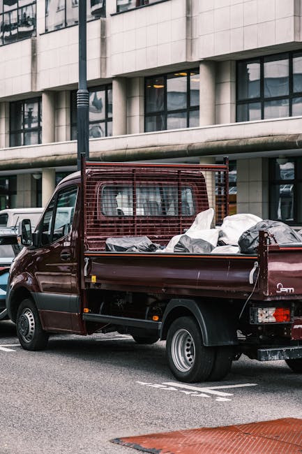 A small burgundy flatbed truck parked on an urban street in front of a modern office building with large glass windows and a white concrete facade. The truck's cargo area is loaded with various types of waste materials, including black and grey plastic bags, some appearing torn or partially open, and loose debris. The flatbed has a low metal side panel and a mesh protective screen at the front of the cargo area, which shows signs of use with some rust and dirt. The vehicle's rear lights are visible, and the truck is positioned in a designated parking zone, with a white van partially visible behind it. Street furniture, such as a pole, is also seen in the background. This scene reflects the type of private waste collection services often used for builder’s rubbish removal in urban settings, carried out by companies like Rubbish Removal Knightsbridge, providing efficient on-site clearance of refuse that may include construction debris, packaging, and other waste materials typical for rubbish removal jobs.
