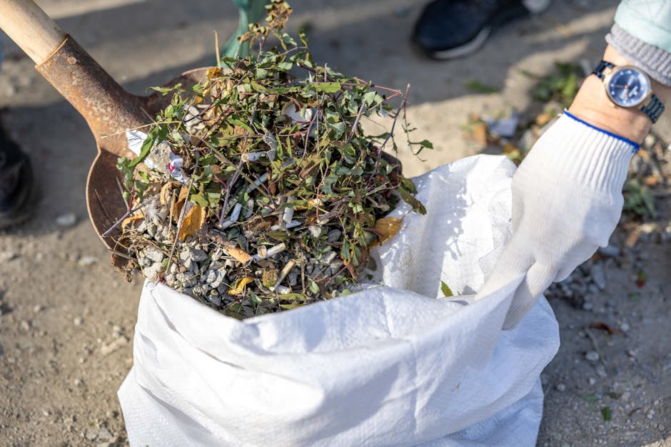A close-up view of a person wearing a white glove with a blue watch on the wrist, holding a large white sack filled with garden waste and debris. The sack contains small branches with green and brown leaves, mixed with gravel, dirt, and small pieces of plant material. A rusty metal shovel with a wooden handle is partially visible, resting against the sack and used for collecting the waste. The background shows a compacted dirt surface with some scattered leaves and debris, indicating an outdoor setting suitable for waste collection or site clearance by a specialist such as Rubbish Removal Knightsbridge. The scene emphasizes on-site greenery disposal, aligning with private, independent waste handling services for rubbish removal in the local area.